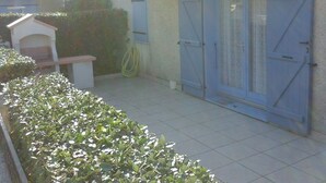 A patio area with a tiled floor, a small white structure, and a hedge.