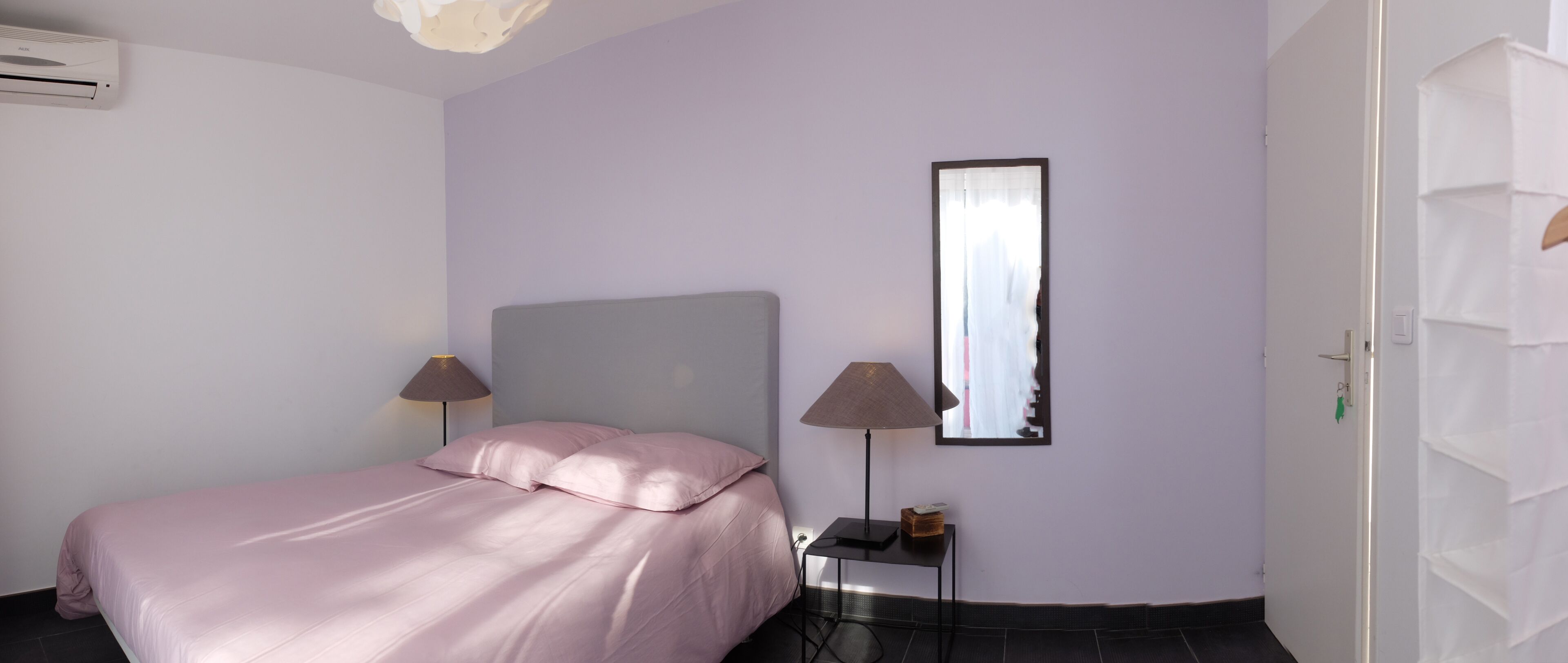 A bedroom with a bed, two bedside lamps, a mirror, and an air conditioning unit.