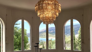 Ein Essbereich mit einem großen Tisch, Stühlen und einer Kronleuchter. Drei Bogenfenster bieten Blick auf eine gebirgige Landschaft.