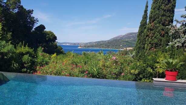 Ein klarer, blauer Infinity-Pool mit einer Holzterrasse, umgeben von üppiger Vegetation und Blick auf das Meer und die Hügel.