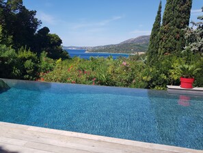 A clear blue infinity pool with a wooden deck, surrounded by lush greenery and a view of the sea and hills. - PROMOTION up to -20% , Magnificent Villa Riviera, sea view, infinity pool, A/C (Le Lavandou)