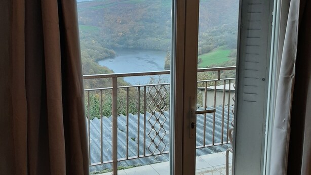 A room with a large sliding glass door leading to a balcony with a metal railing and a view of a valley and a lake. - Randonnée portes de l'Aubrac Vue sur Lac 4 pers 2 chambres 2wc Mandailles (Castelnau-De-Mandailles)