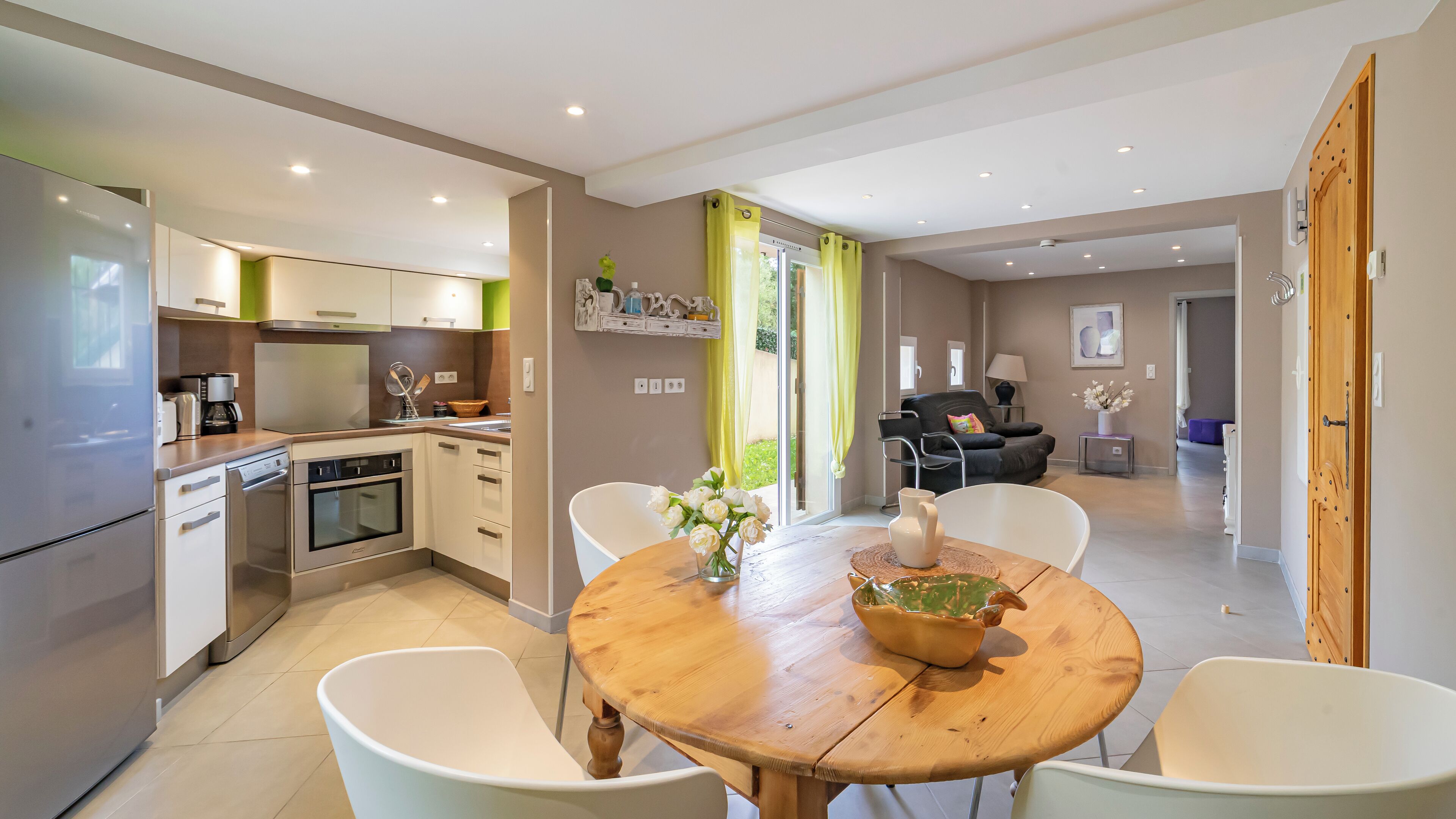 Een moderne keuken met witte kastjes, een ingebouwde oven en een koffiezetapparaat. Er is een eethoek met een houten tafel en witte stoelen.