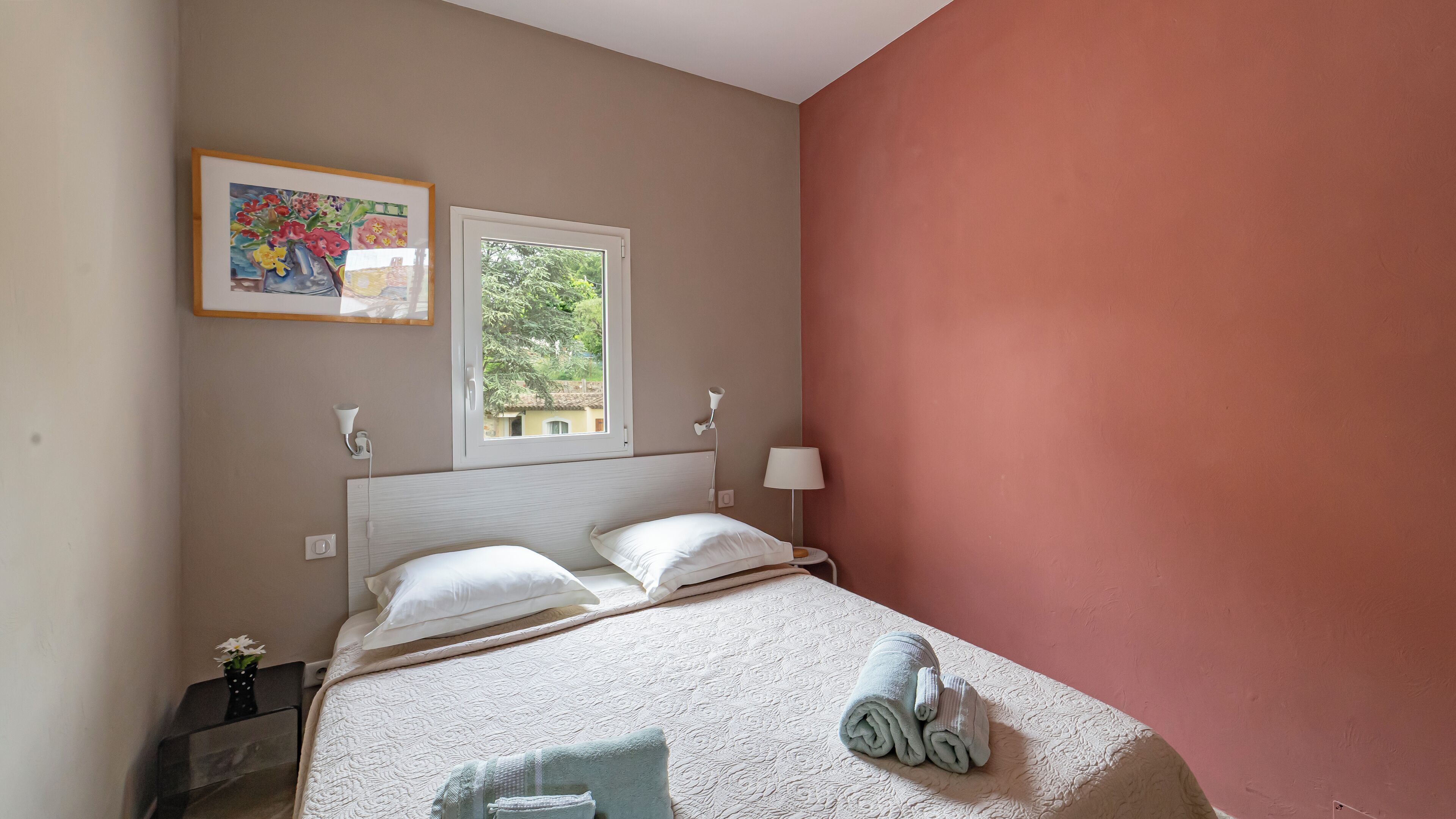 Ein Schlafzimmer mit Bett, Kissen, einer Nachttischlampe, einem Fenster und einem Bild an der Wand.