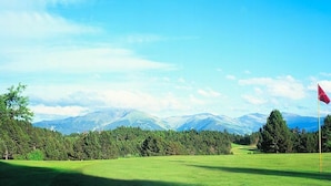 A golf course with a red flag, surrounded by trees and mountains in the background.