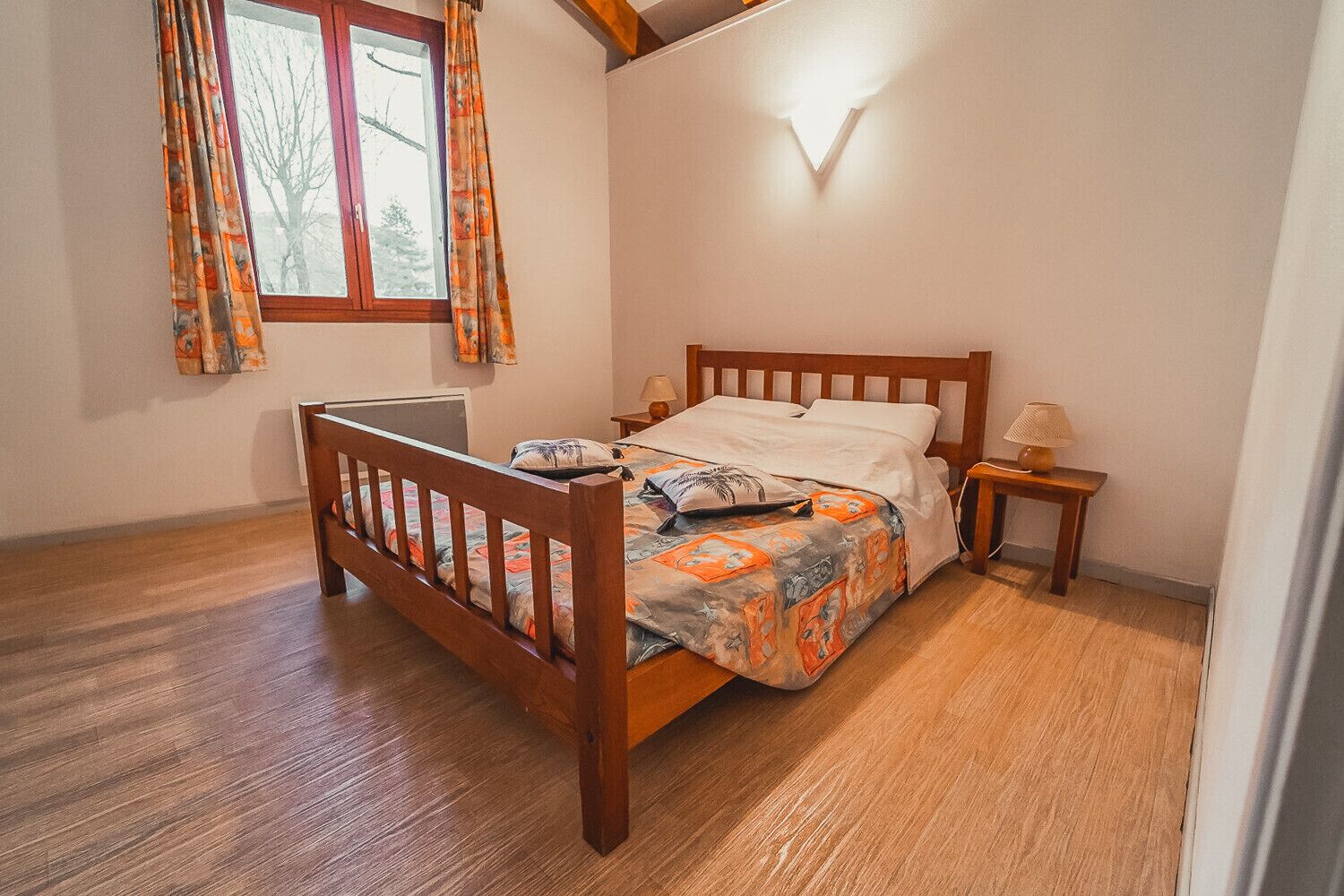 Ein Schlafzimmer mit einem Holzbett, einem Nachttisch mit Lampe und einem Fenster mit Vorhängen.
