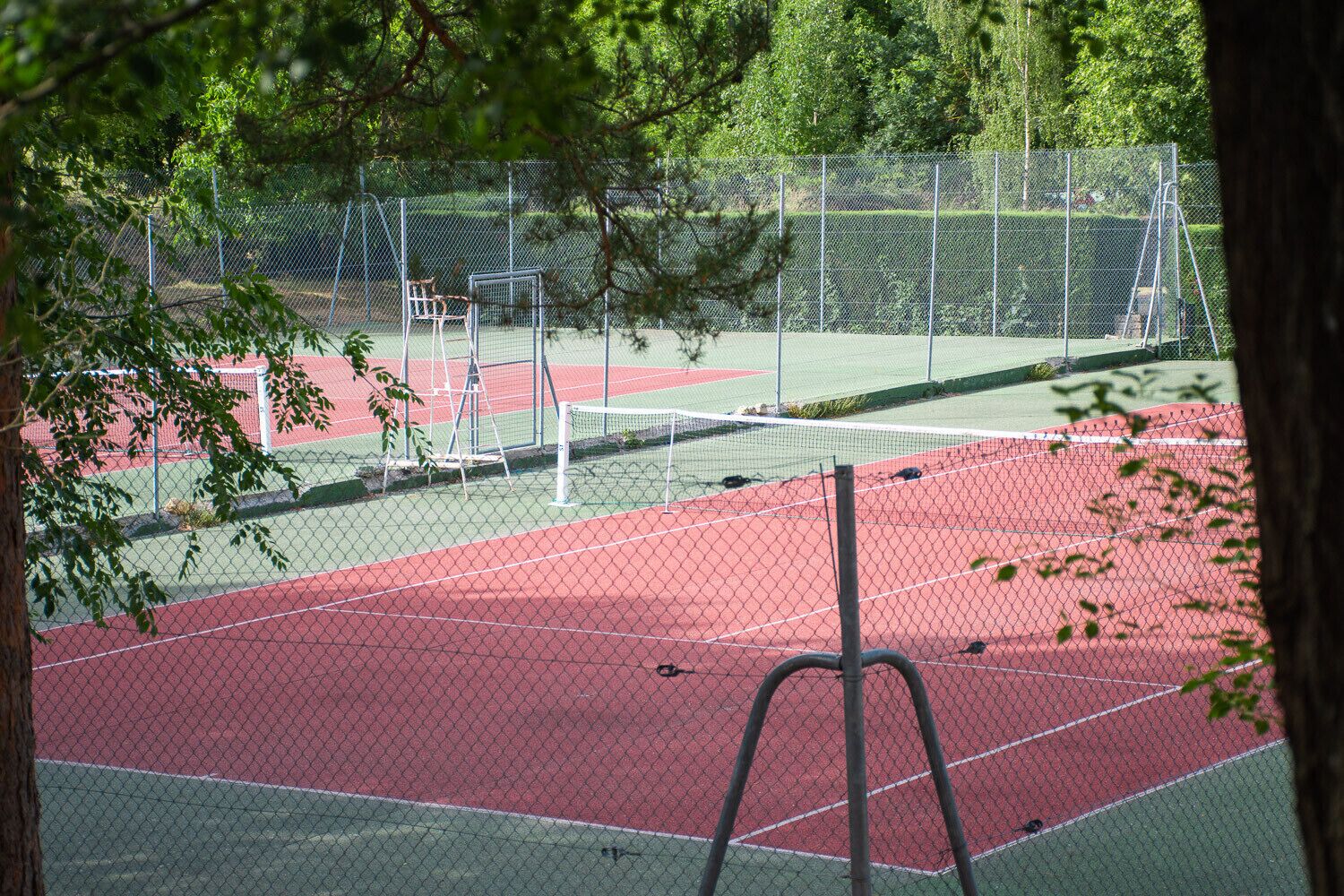 Ein Tennisplatz im Freien mit roter Belag, umgeben von einem Maschendrahtzaun und Bäumen.