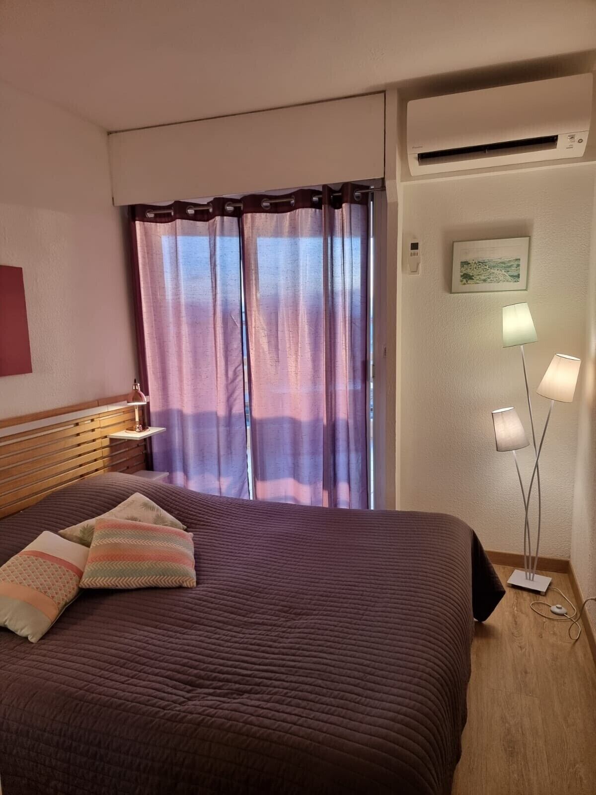 A hotel room with a bed, curtains, a bedside lamp, an air conditioning unit, and a wall-mounted picture.