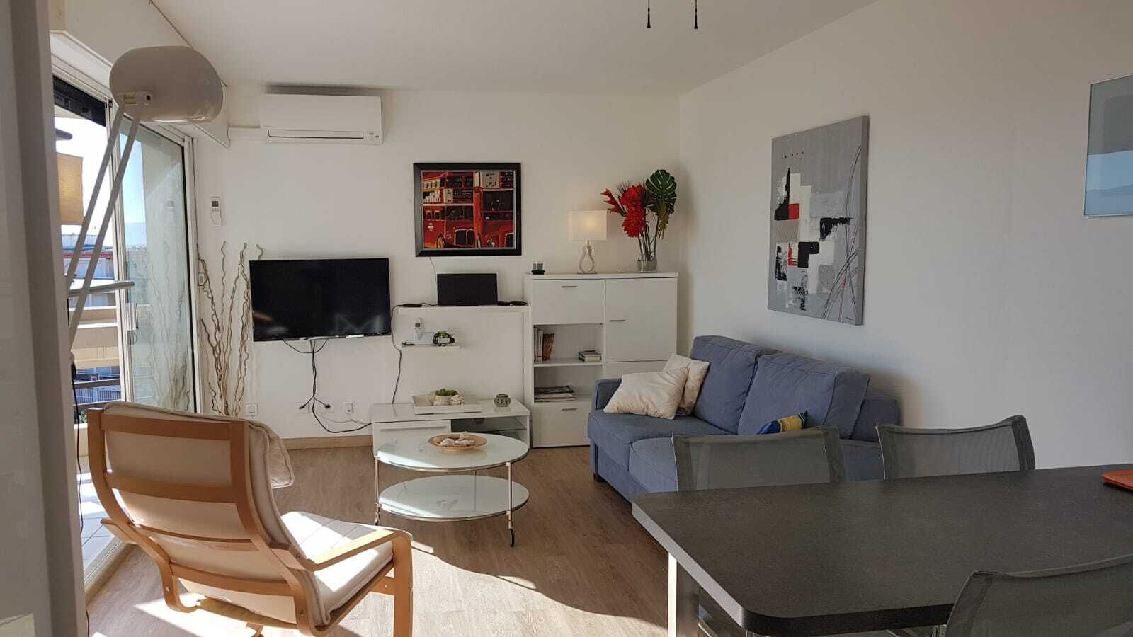 A modern living room with a sofa, coffee table, and a dining table. There is a TV mounted on the wall, a small white cabinet, and a plant on the windowsill.