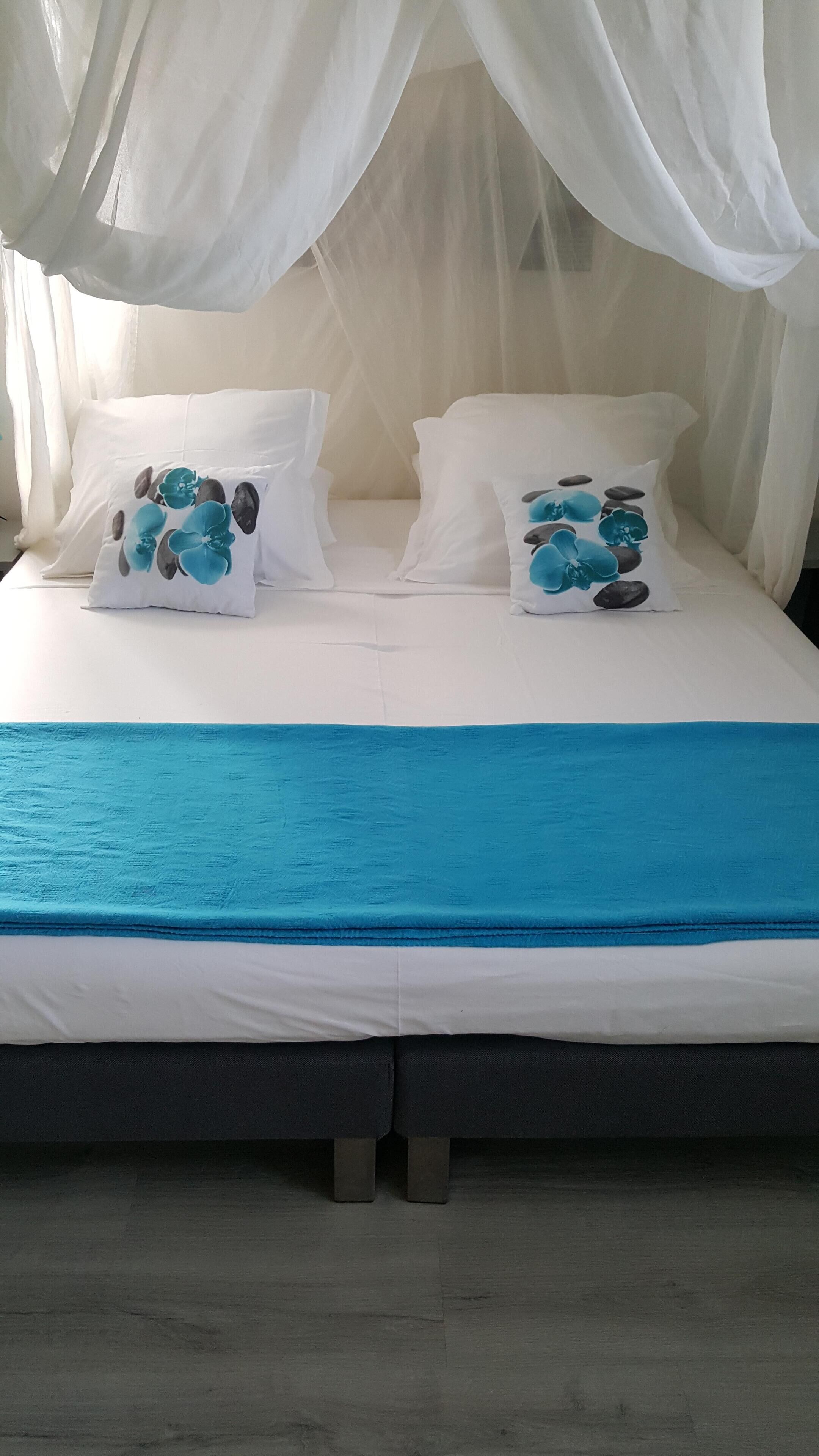 Ein Bett mit weißer Bettwäsche, zwei dekorative Kissen mit blauen und grauen Mustern, eine blaue Decke und ein weißes Moskitonetz.