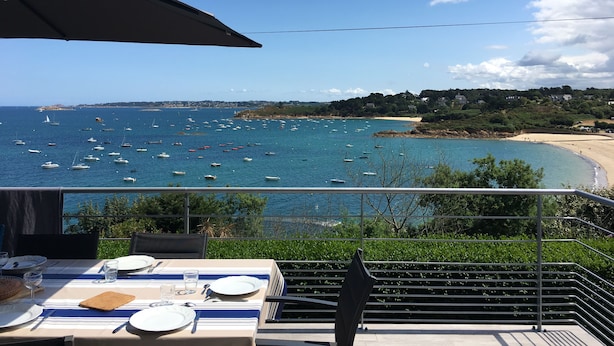 A dining table set with plates, cutlery, and glasses, overlooking a coastal view with boats and a beach. - Breton family house with magnificent sea views (Carantec)