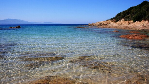 A clear coastal beach with rocky terrain and a distant mountain range.