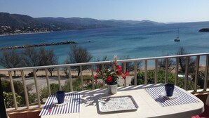Un balcon avec une table et des chaises, offrant une vue sur la côte, avec un ciel dégagé et des montagnes lointaines.