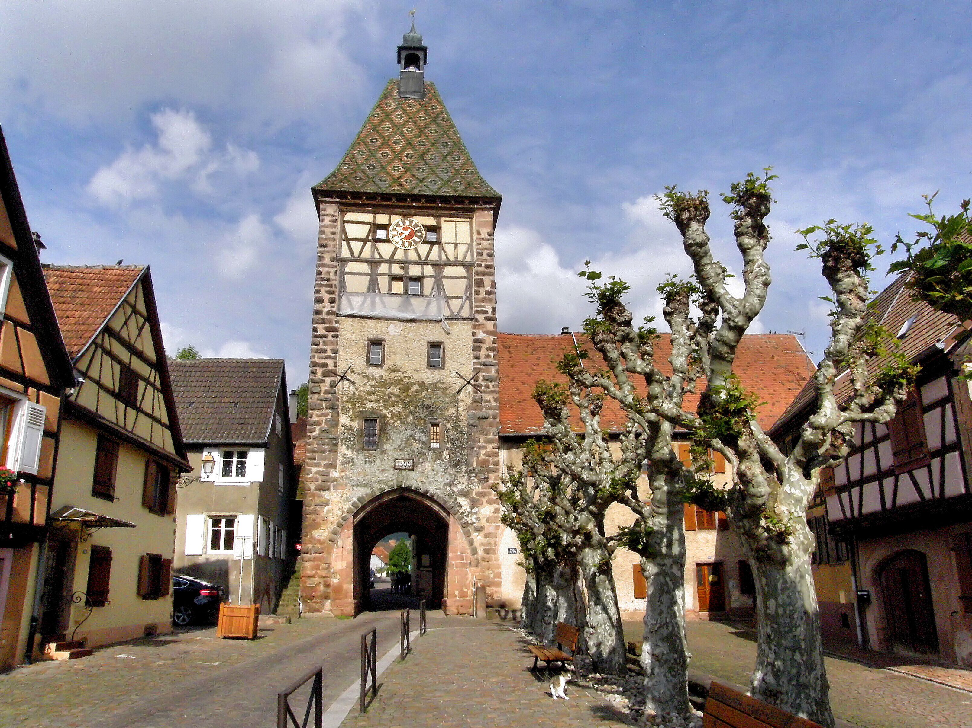 Ein historischer SteinTurm mit einer Uhr, umgeben von traditionellen Fachwerkhäusern und einer Kopfsteinpflasterstraße.
