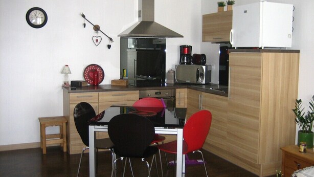 A compact kitchen with a microwave, toaster, and coffee maker. There is a dining table with chairs and a small stool. The kitchen has a wooden cabinet and a white refrigerator.