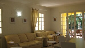 A living room with a beige sofa, a small round table, and a dining table with chairs. There are two windows with curtains, and artwork on the walls.