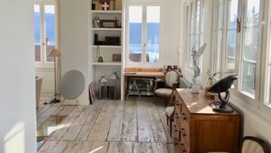 Une pièce dotée d’un plancher en bois, d’un bureau avec une chaise, d’une bibliothèque et offrant une vue sur la mer grâce à de grandes fenêtres.