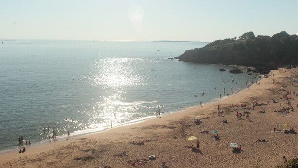 Ein Strand mit badenden Menschen, einer felsigen Küste und blauem Himmel.