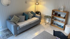 Ein Wohnzimmer mit grauem Sofa, gemusterten Kissen, einem kleinen runden Tisch, einem Bücherregal und einer Stehlampe.