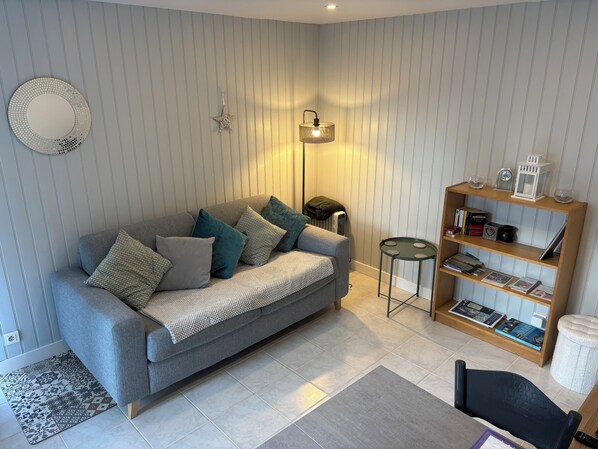 A living room with a grey sofa, patterned cushions, a small round table, a bookshelf, and a floor lamp.