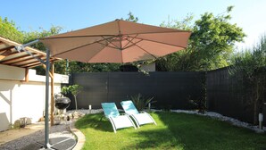 Ein Garten mit einer Terrassenfläche, zwei Liegestühlen, einem Grill und einem großen Sonnenschirm.