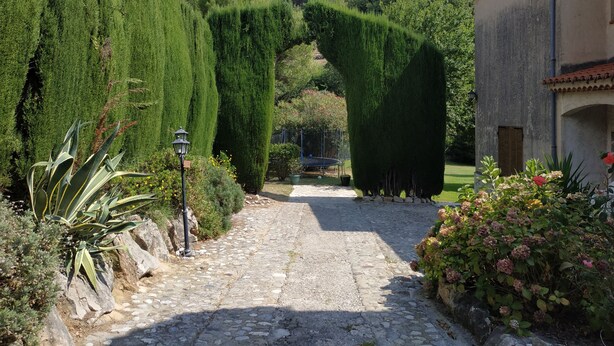 Ein Kopfsteinpflasterweg schlängelt sich durch einen Garten mit ordentlich geschnittenen Hecken und ein Steinhaus mit Ziegeldach.
