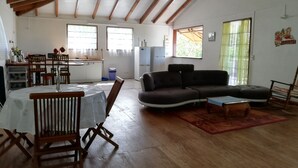 A spacious living room with a wooden ceiling, a dark sofa, a dining table with chairs, and a kitchen area in the background. - Maisoson 130m in Sainte-Rose for the holidays at the seaside, mountains. (Sainte-Rose)