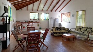 A spacious living area with wooden flooring, a dining table with chairs, a sofa, and a rocking chair. There are windows with curtains, a refrigerator, and a television on a stand. - Maisoson 130m in Sainte-Rose for the holidays at the seaside, mountains. (Sainte-Rose)