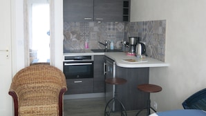 Cocina compacta con armarios oscuros, salpicadero de azulejos, horno empotrado y cafetera. Hay dos taburetes y una silla de mimbre.