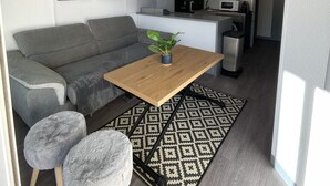 A compact living space with a grey sofa, a wooden coffee table with a plant, two grey poufs, a patterned rug, and a kitchenette in the background. - Les Goëlettes apartment in Saint Cyprien Plage, Pyrenees - Orientales - 4 people (Saint-Cyprien)