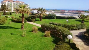 A well-maintained garden with a pathway, palm trees, and a building with a red roof in the background. - Les Goëlettes apartment in Saint Cyprien Plage, Pyrenees - Orientales - 4 people (Saint-Cyprien)