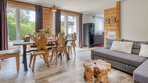 Una sala moderna con un sofá gris, una mesa de comedor de madera con sillas y una zona de cocina con refrigerador.