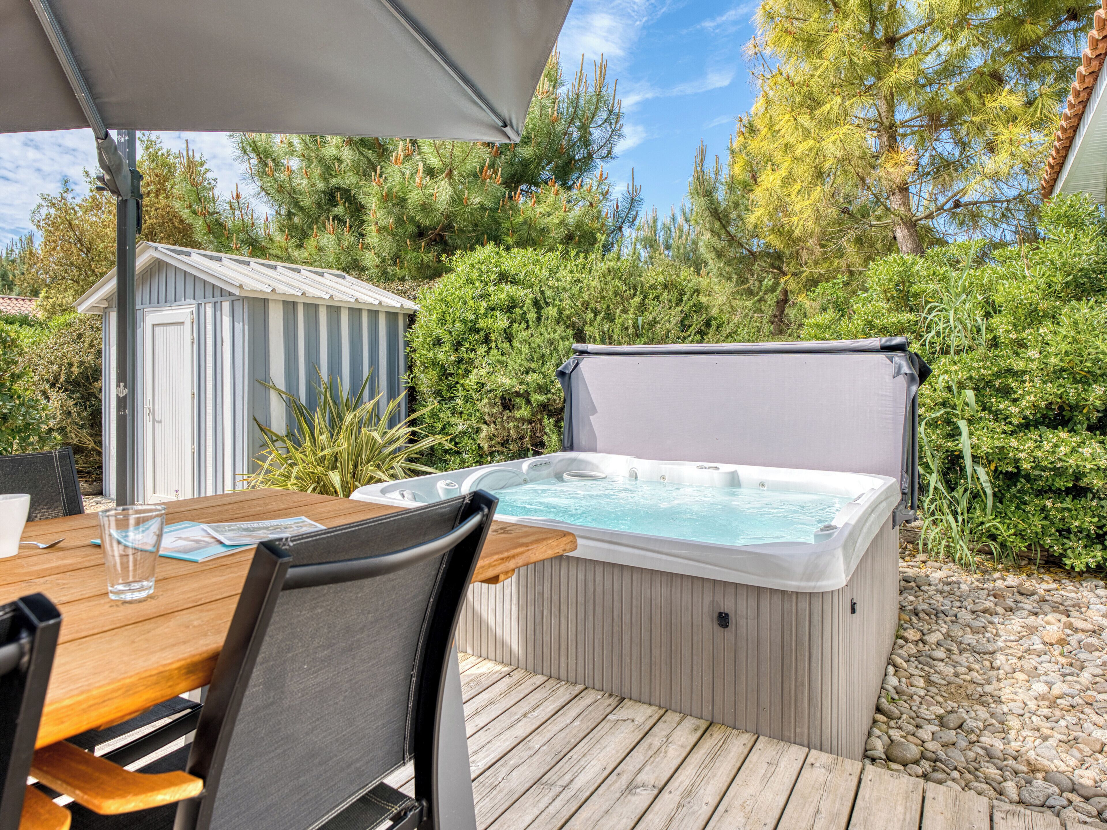 A hot tub on a wooden deck with a table and chairs, surrounded by greenery and a shed.