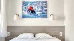 Una cama con sábanas blancas, cabecera de madera, dos lámparas fijadas a la pared e imagen con motivos invernales en la pared.