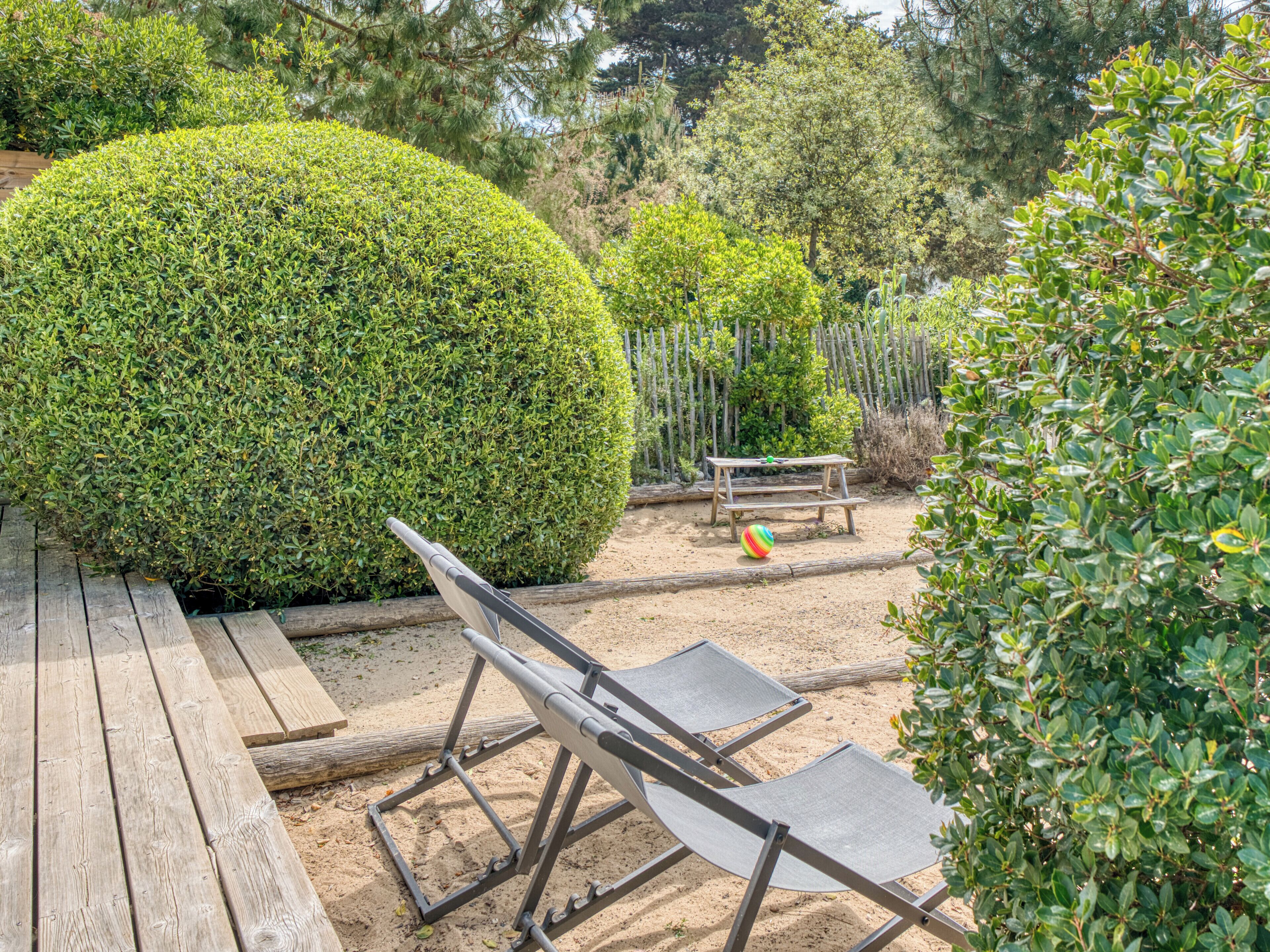 Una terraza de madera con una silla de metal y un banco en un jardín con arbustos y árboles bien cuidados.