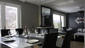 Un espace repas moderne avec une table blanche dressée pour six personnes, entourée de chaises noires. Un grand miroir reflète la pièce, et un canapé gris est visible au fond.