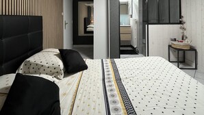 Une chambre à coucher moderne avec un lit, une table de chevet, un miroir et une salle de bain visible à travers une porte ouverte.