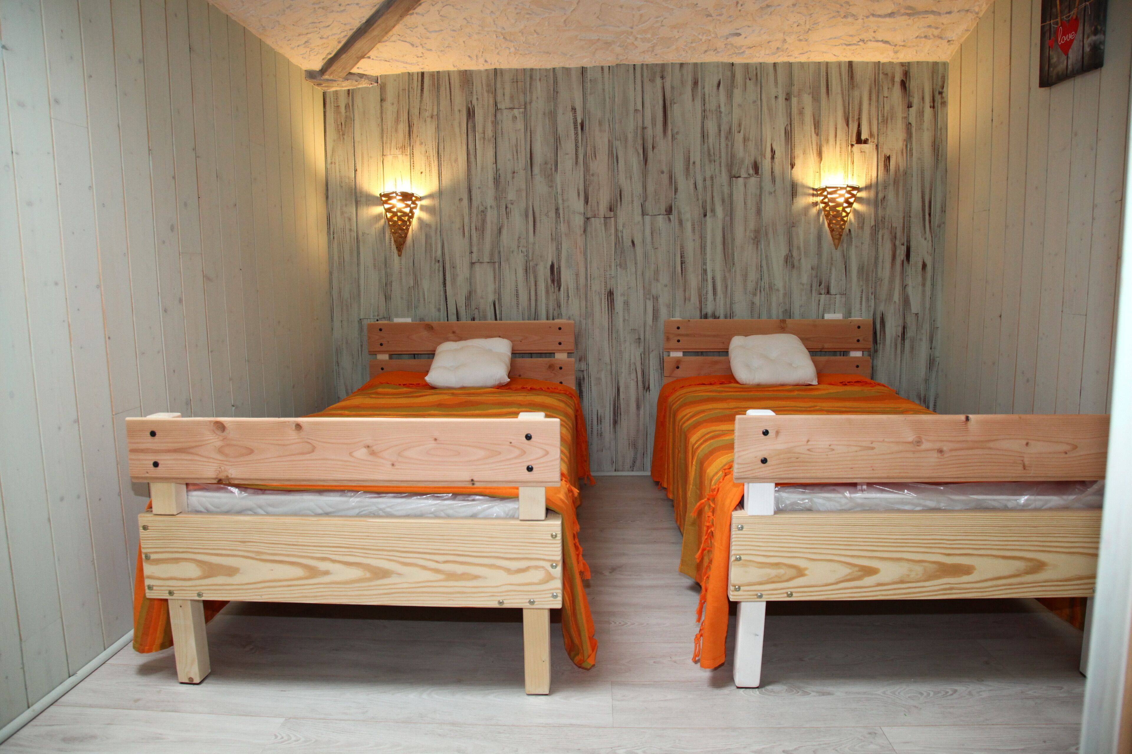 Une chambre avec deux lits en bois, chacun étant muni d’un oreiller et d’une couverture orange.