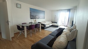 Un salon moderne avec un canapé, une table à manger et des chaises, un tableau représentant un paysage urbain et une grande fenêtre avec des rideaux.
