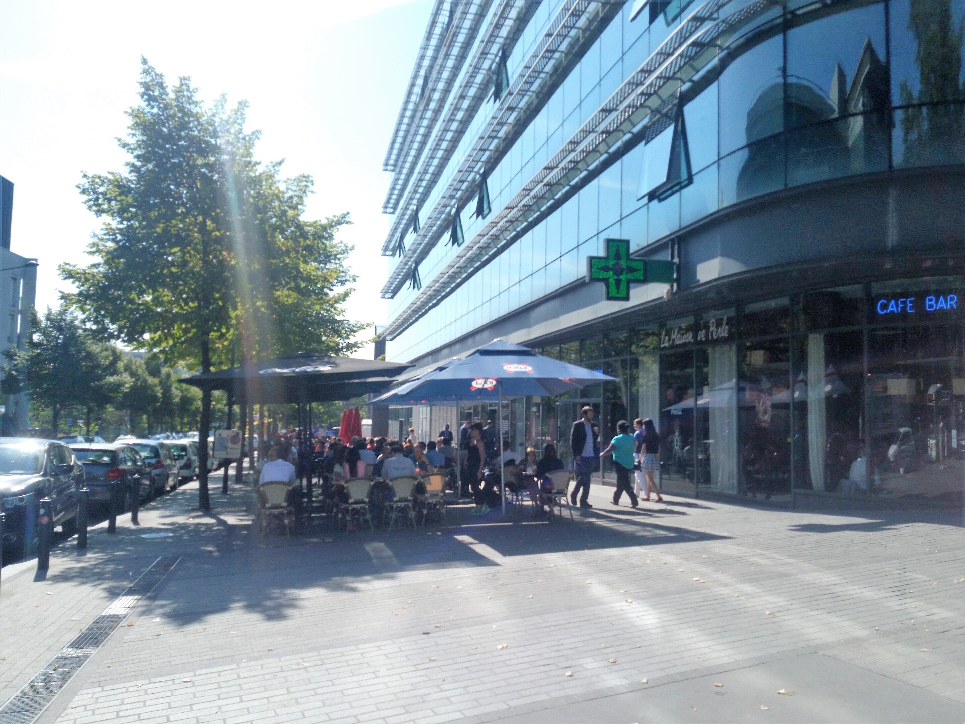 An outdoor cafe with tables and umbrellas, a cafe bar, and a building with a green cross sign.