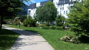 Un chemin mène à un bâtiment moderne à plusieurs étages, entouré de pelouses et d’arbres bien entretenus.