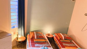 Une petite chambre agréable, dotée de deux lits, d’une table de chevet, d’une lampe et d’une fenêtre avec des stores.