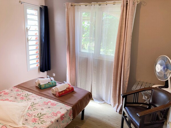 A room with a bed, a chair, a fan, and a window with curtains.