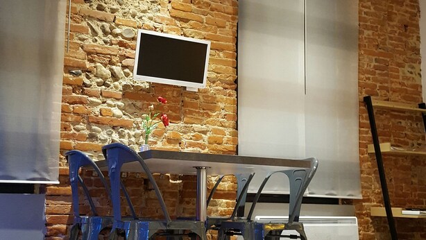 A dining area with a brick wall, a table, chairs, a TV mounted on the wall, and a window with blinds. - Toulouse Canal du Midi (Toulouse)