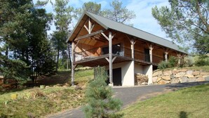 A two-story log cabin with a balcony, surrounded by trees and a stone wall.