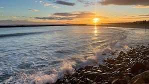 A beach scene with the sun setting over the horizon, waves crashing onto the shore, and a rocky coastline.