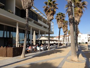An outdoor seating area with palm trees and umbrellas, a modern building with balconies, and a clear sky.