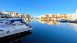 A marina with boats docked, a clear blue sky, and buildings with balconies and palm trees.