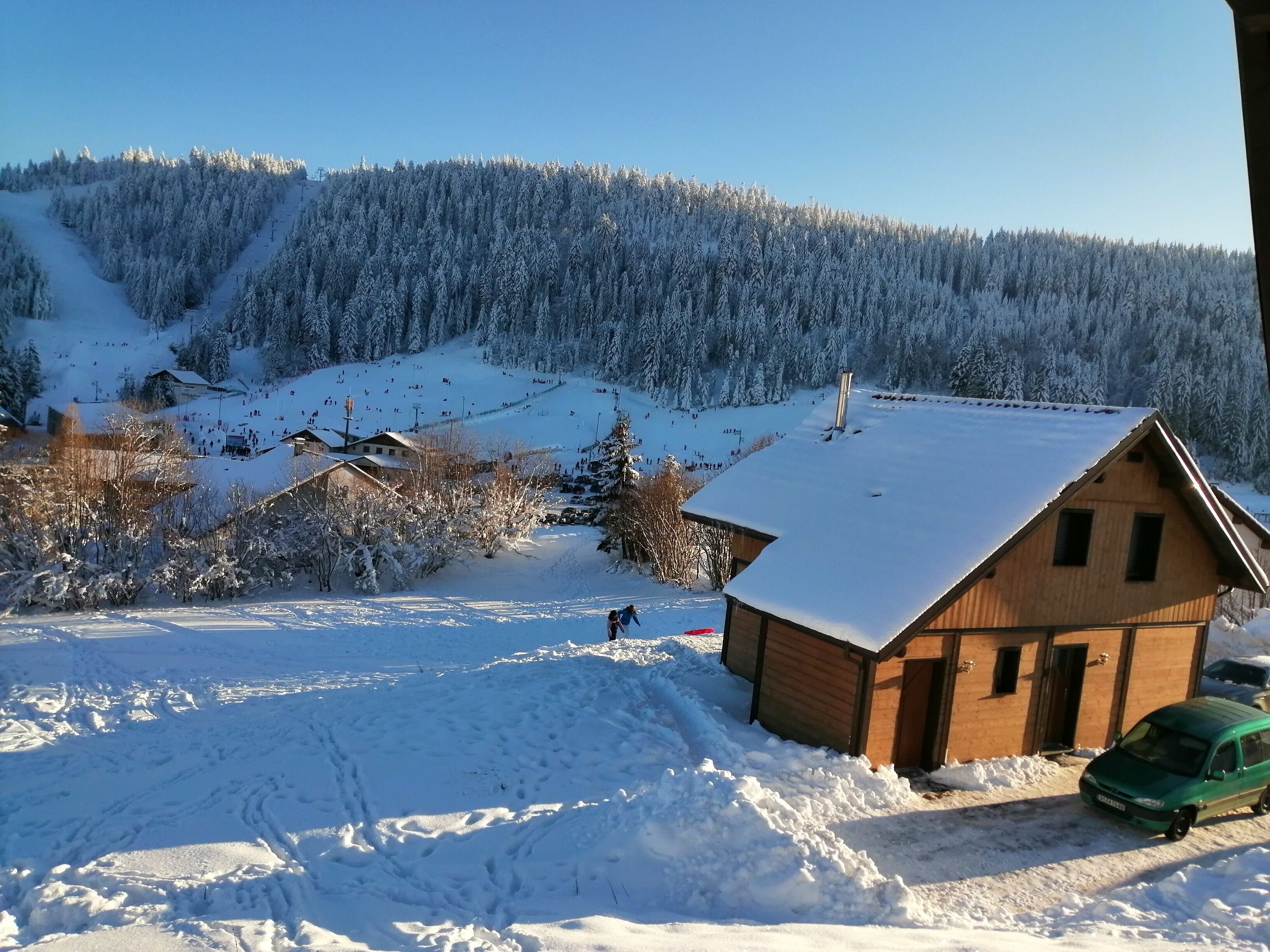 Een houten hut met een met sneeuw bedekte dak, omgeven door een besneeerd landschap en een bebost heuveltje op de achtergrond.