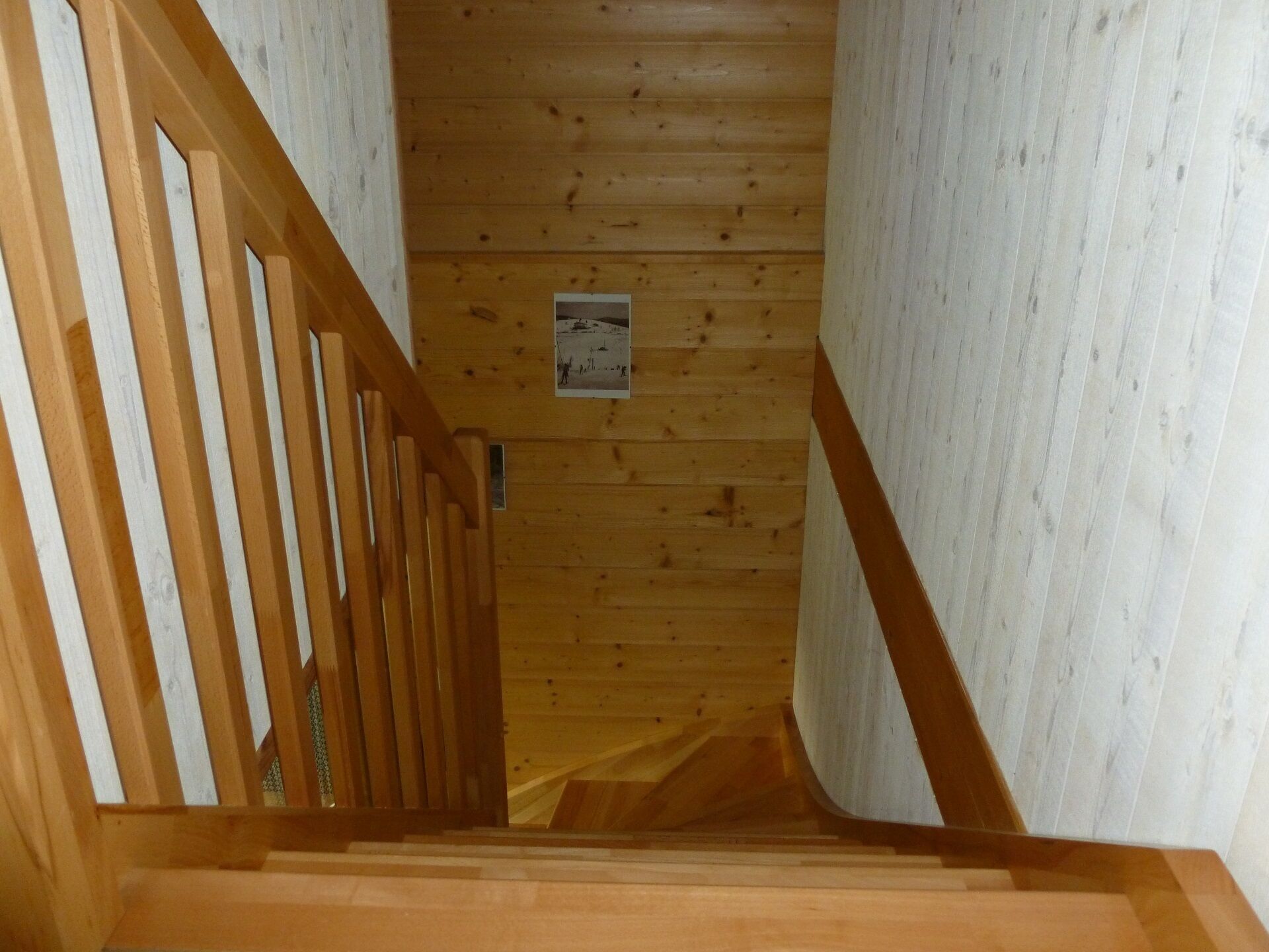 Un escalier en bois avec une rampe, menant à une pièce dont un mur est blanc et qui possède une petite fenêtre.